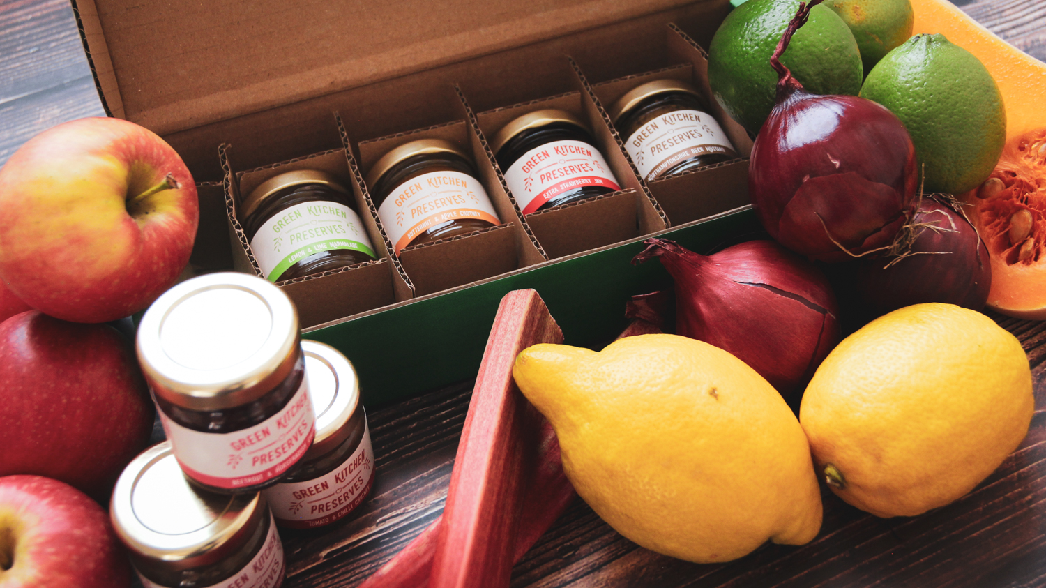 an assorted box of mini jars of chutneys, jams and marmalades on a wooden background with fruit and vegetables around it