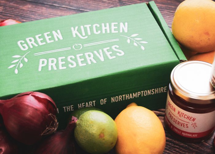 a green kitchen preserves gift box of jars containing jams, chutneys and marmalades on a wooden background with fruit and vegetables on a wooden background