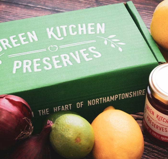 a green kitchen preserves gift box of jars containing jams, chutneys and marmalades on a wooden background with fruit and vegetables on a wooden background
